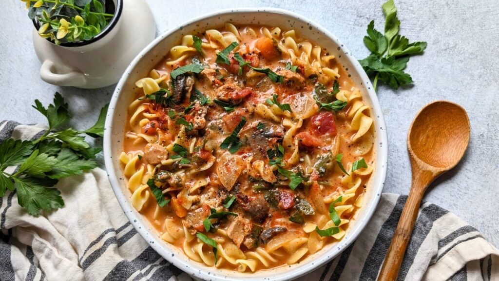 hungarian soup recipe with chicken, spicy paprika, onions, mushrooms, tomatoes, and fresh chopped parsley over egg noodles in a white bowl with a wooden spoon