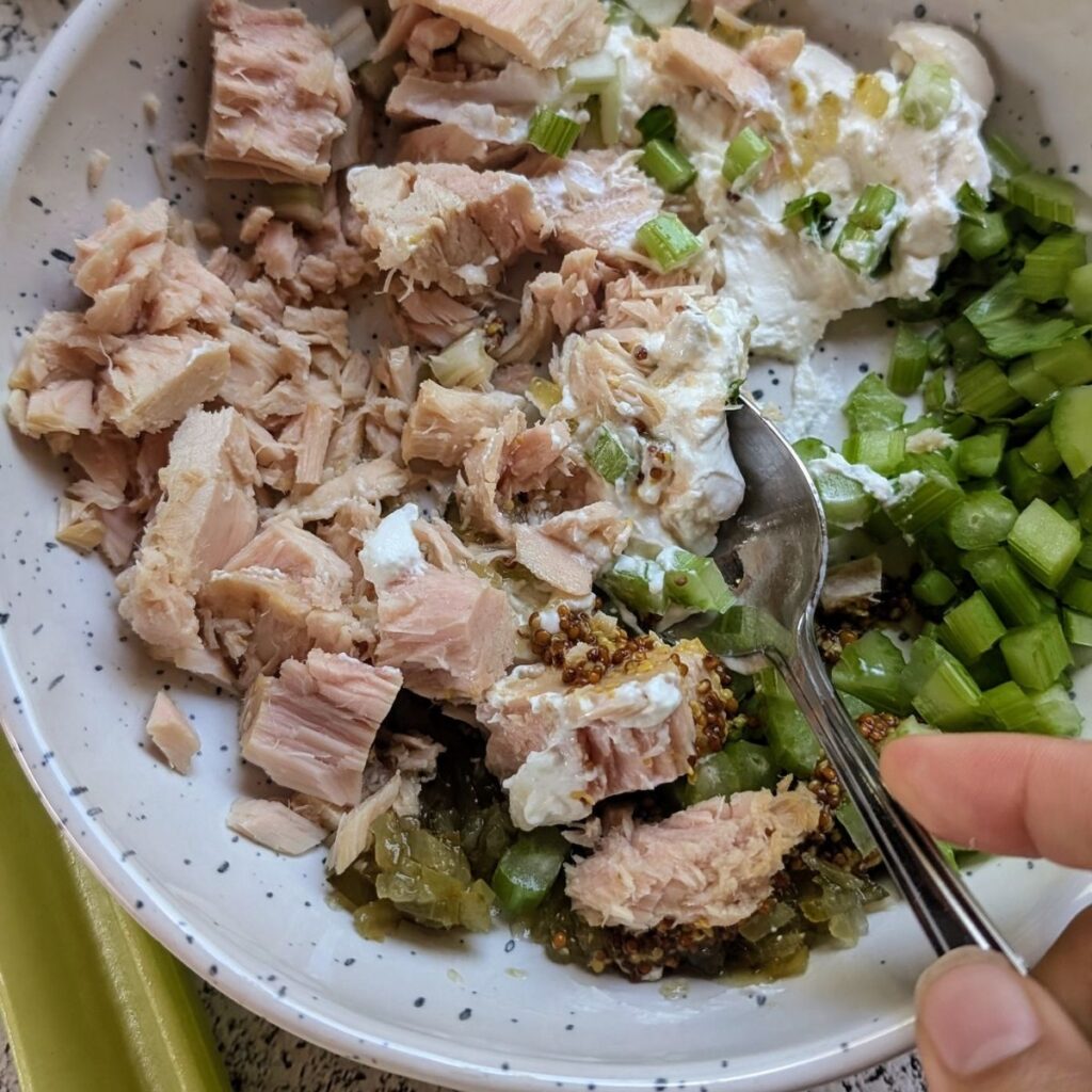 a spoon mashing chunk tuna into tuna salad with Greek yogurt and vegetables for a quick and easy lunch.