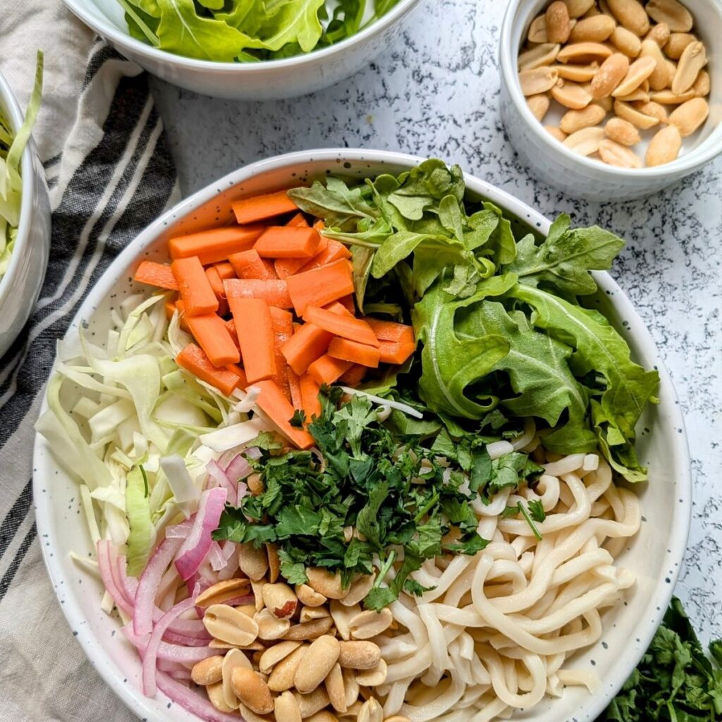 a bowl full of thai wheat noodles, carrots, cilantro, pickled onions, cabbage, and fresh arugula.