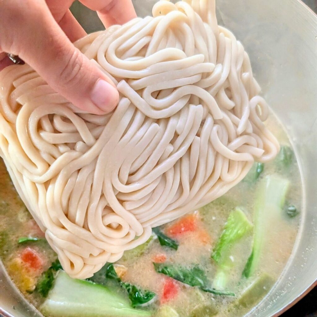 adding thai wheat noodles to a hot pot of soup with thai basil, lime, coconut milk, and soy sauce.