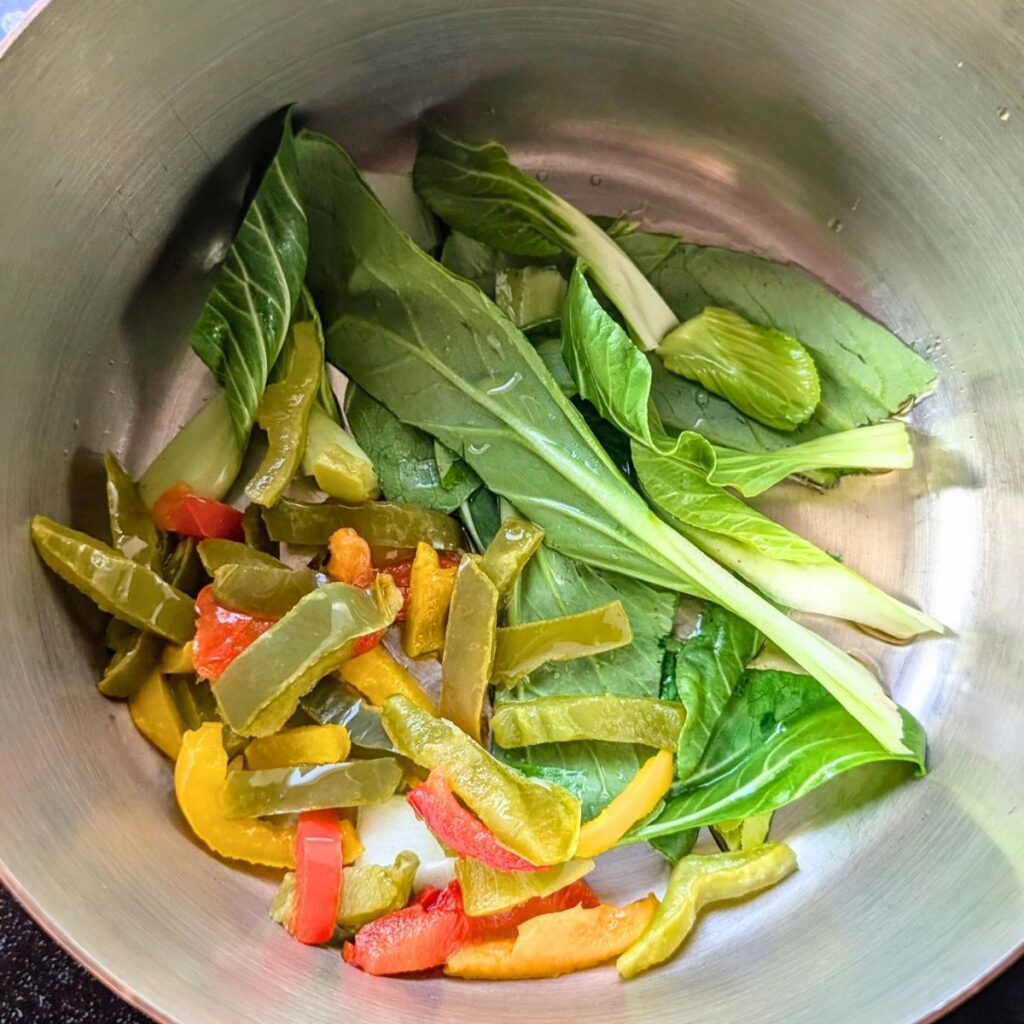 a pot with bok choy, garlic, and bell pepper strips sauteeing to make thai wheat noodle soup.