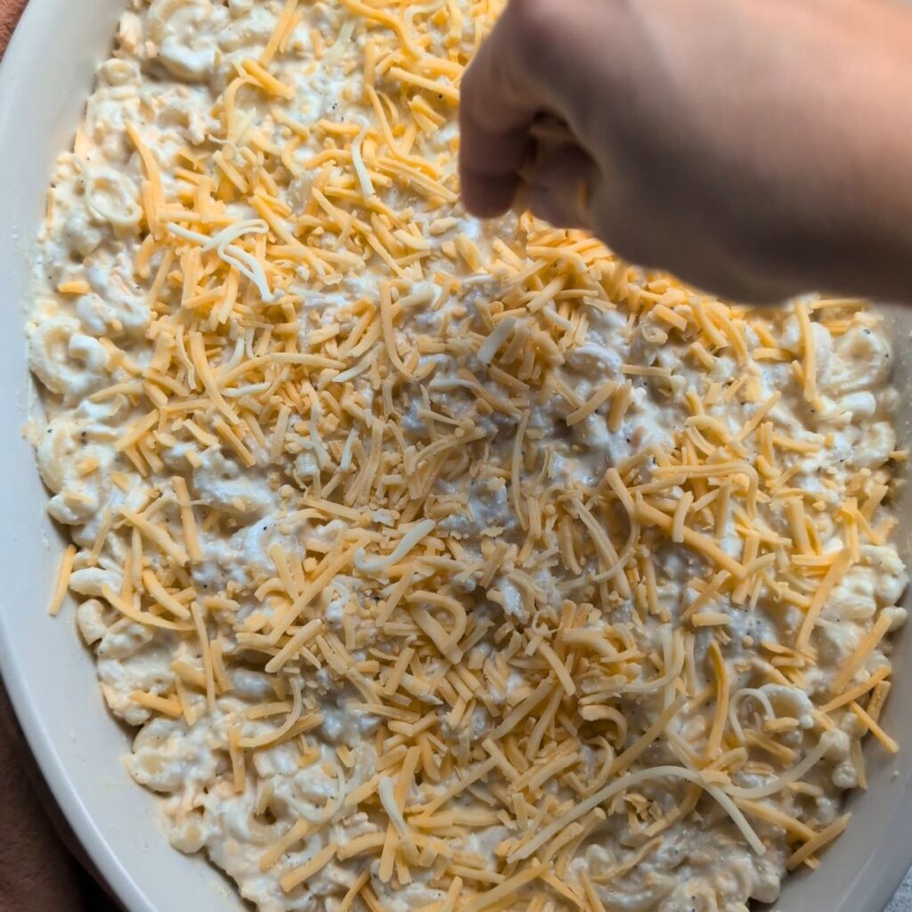 a hand sprinkling cheddar cheese on top of creamy ricotta cheese macaroni and cheese in a baking dish.