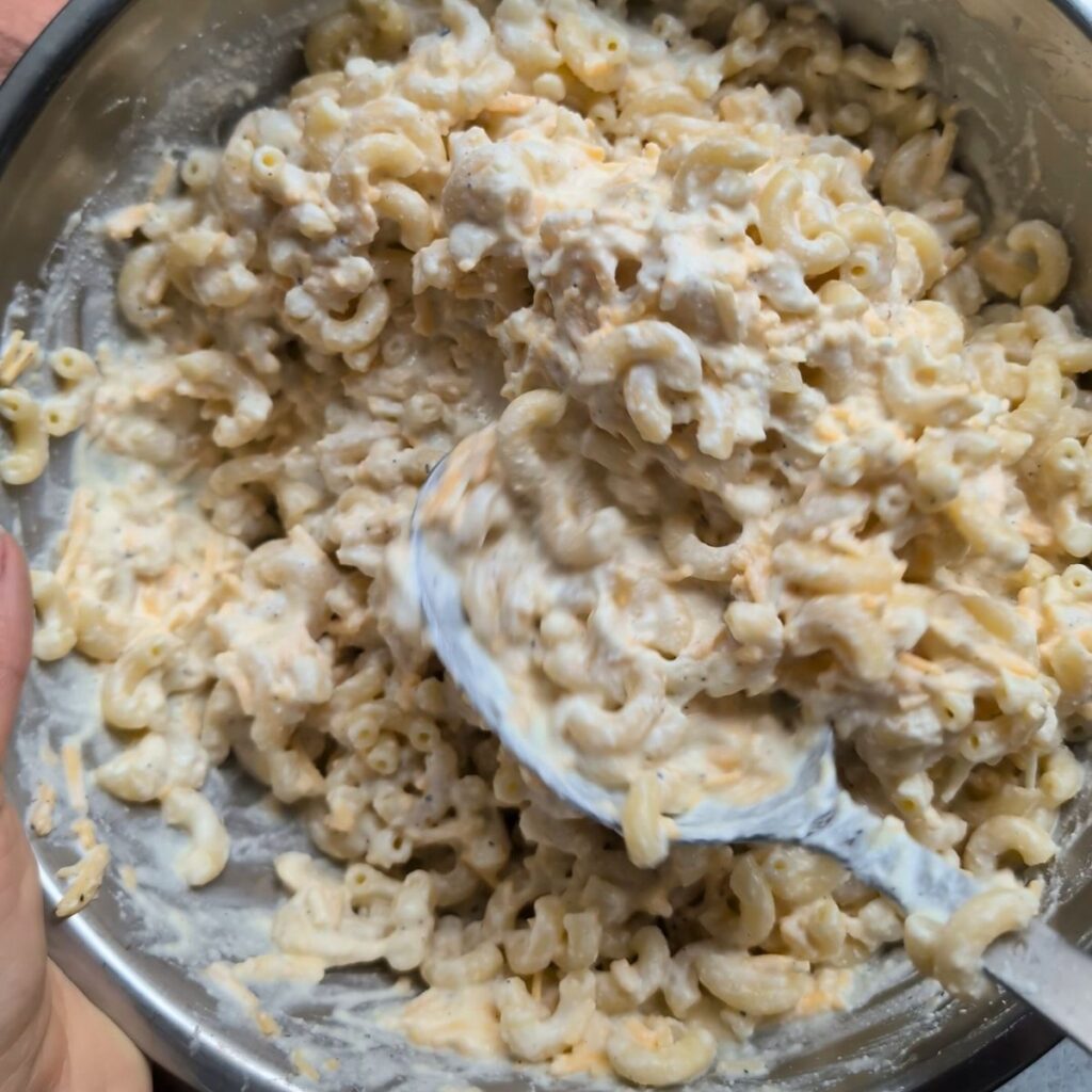 a spoon stirring a large bowl of macaroni and cheese with ricotta, sour cream, spices, and pepper.