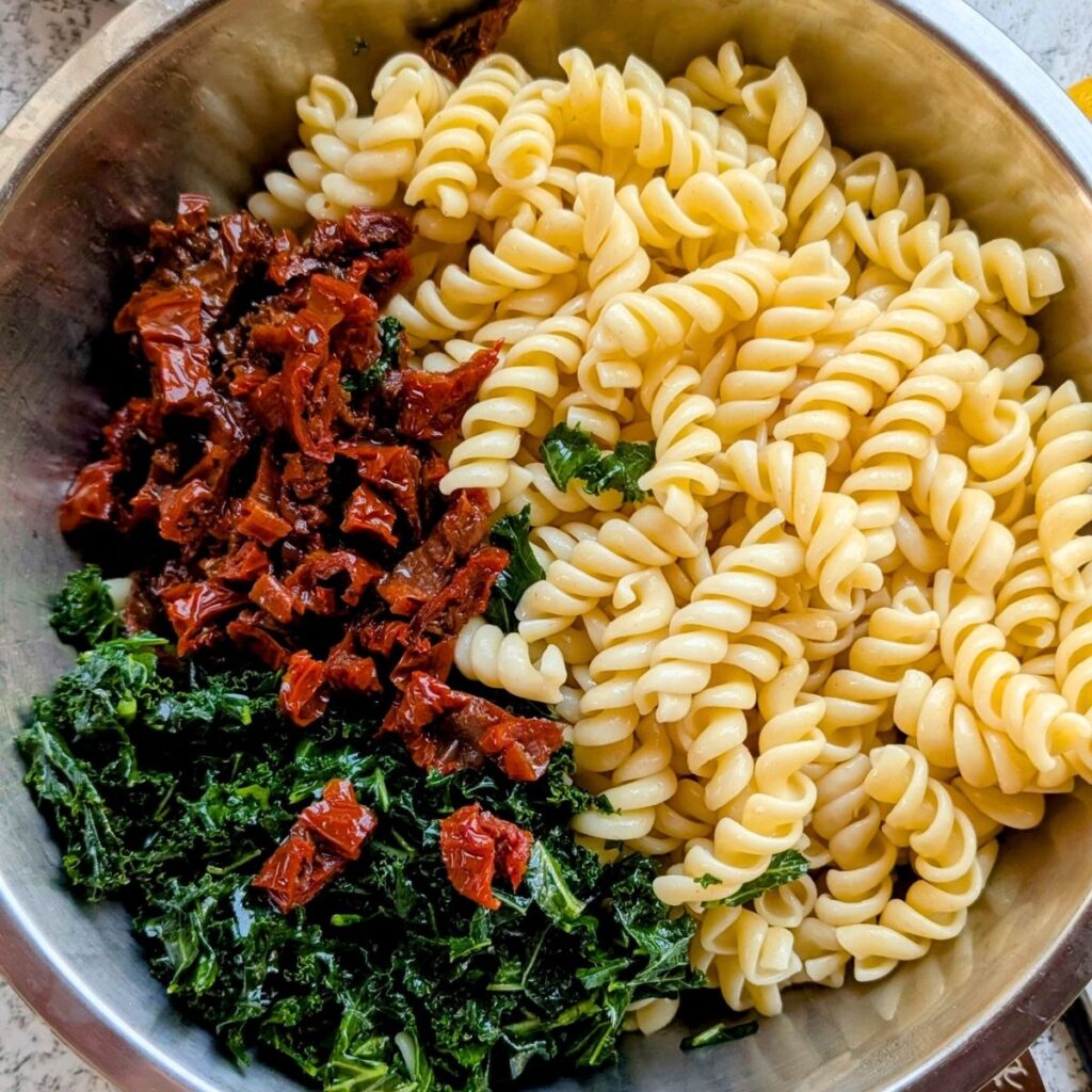 a large bowl with cooked rotini pasta, chopped sun-dried tomatoes, and massaged kale.