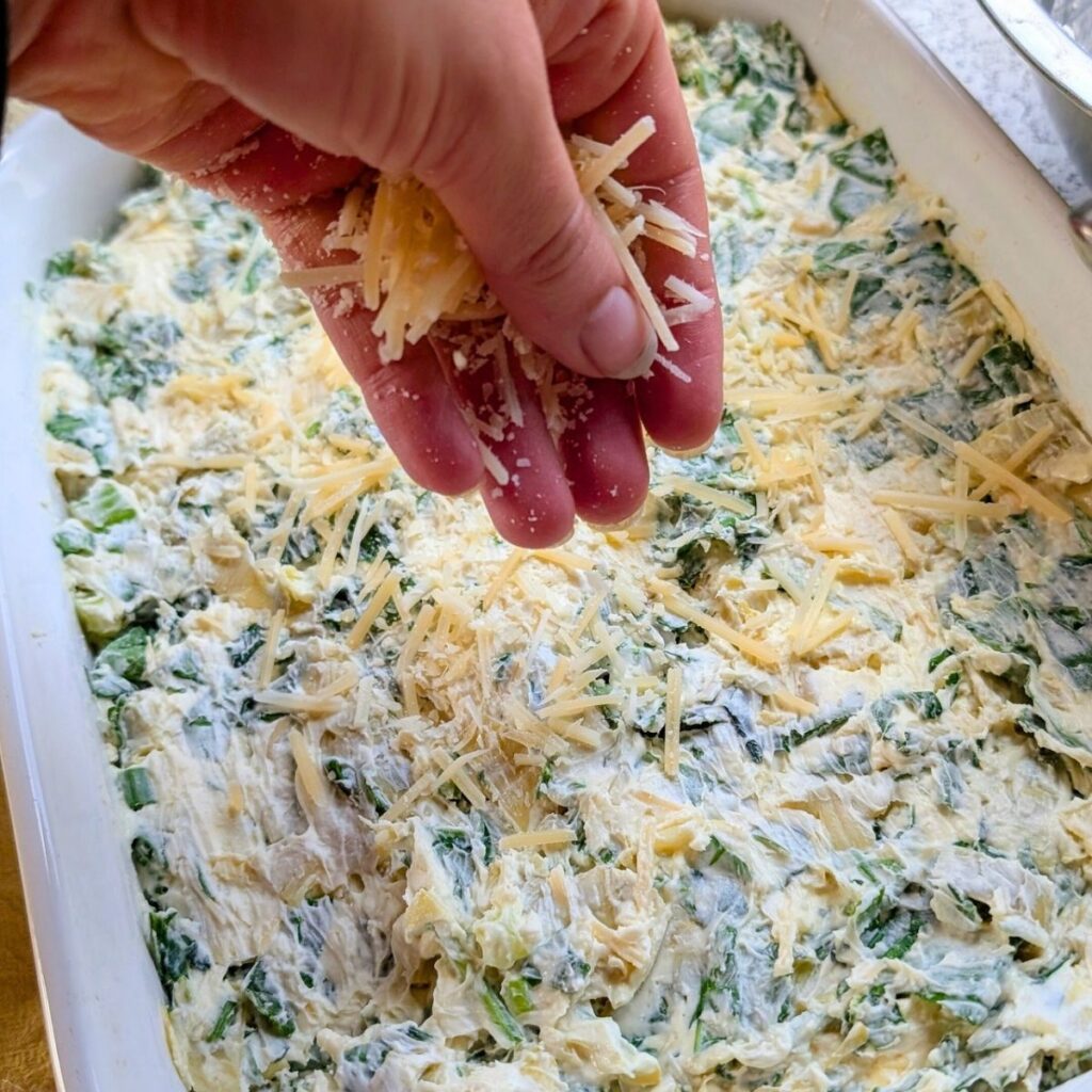 a hand sprinkling parmesan cheese over italian spinach dip in a baking dish.