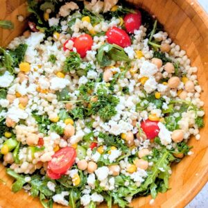 a bowl of couscous salad with arugula and corn and cherry tomatoes in a bowl with feta cheese.