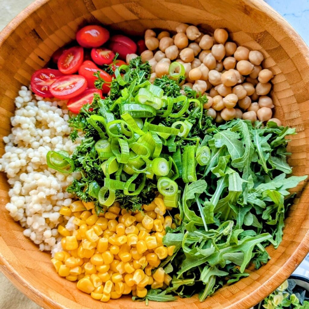 a large salad bowl with couscous, corn, arugula, tomatoes, green onions, and chickpeas.
